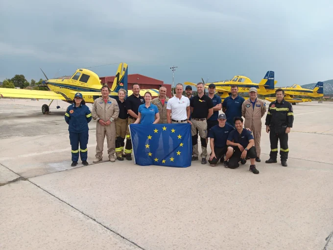 A group picture of firefighters holding the EU flag and standing in front of parked firefighting airplanes.