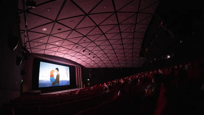 An audience sits in a large cinema auditorium facing a screen at the front showing an animation of two people embracing