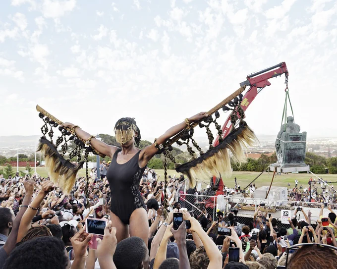 A person wearing a mask and sleeves made from feathers stands above a crowd of people as a statue is being removed.