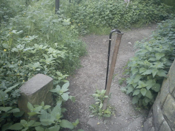 Picture shows a walking stick hanging on a piece of metal pipe standing in the middle of a narrow path. The pipe has a rough, jagged edge at the top, and blocks access to mobility vehicles. Obstructions like this on public rights of way are illegal, but nonetheless extremely common.