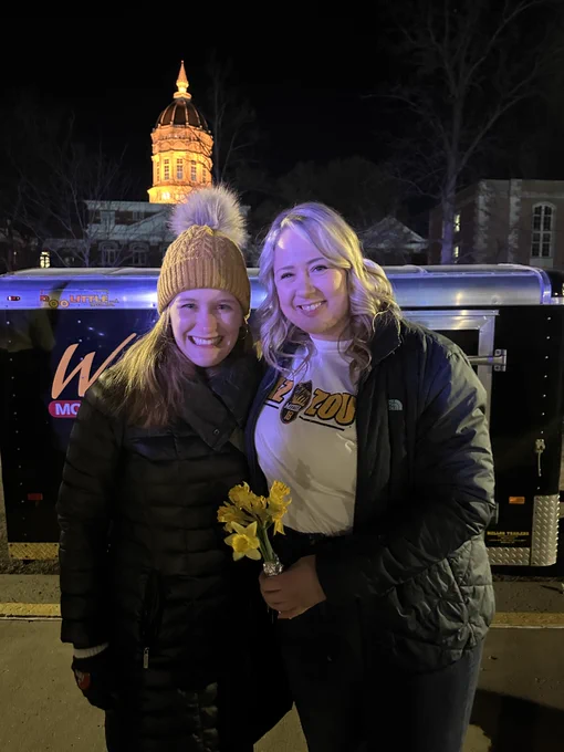 Dr. Alaina Leverenz is posing with her advisor and mentor Dr. Haley Horstman with a gold-glowing Jesse Hall in the background. Haley has a gold beanie on and Alaina has a white Mizzou 18 shirt on. She is holding flowers.