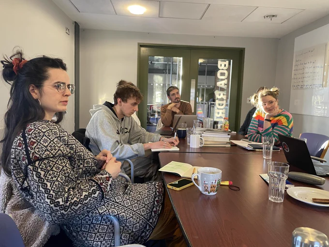 4 young white people sat around a large table listening to someone speak (unpictured)