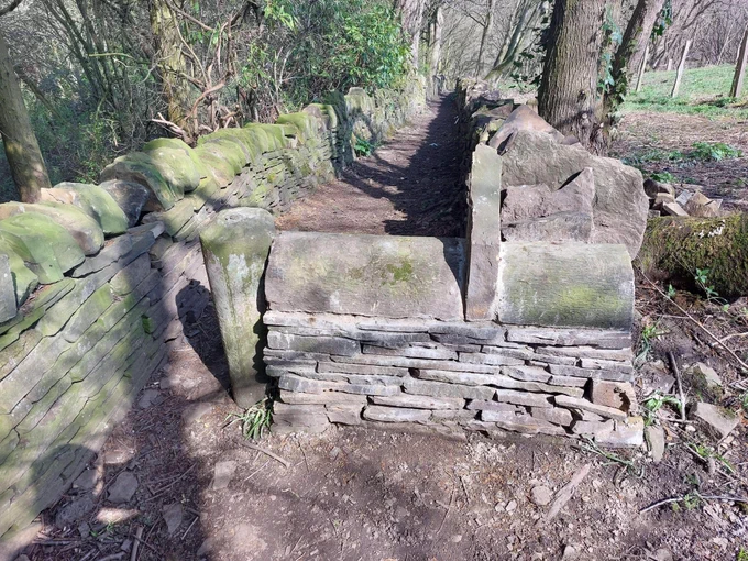 Image shows a wide, level public footpath with a stone wall built across it leaving a gap of about 300mm to squeeze through. There is no legal justification for a structure at this point on the public right of way, and even if there were, structures have been legally required to take into account the needs of mobility-impaired people and permit mobility vehicles for decades. Obstructing a public footpath in this manner is a criminal offence, but sadly very little is done to enforce this.