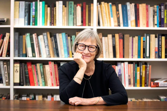 Color photograph of an older white woman. She sits at a wooden table with one arm resting on the table, the other is propping her head up at her chin. She had chin-length gray hair, wears black-rimmed glasses and is wearing a black jacket with a black shirt and a necklace. Behind her is a wall to wall bookshelf with many different colored books on the shelves. 