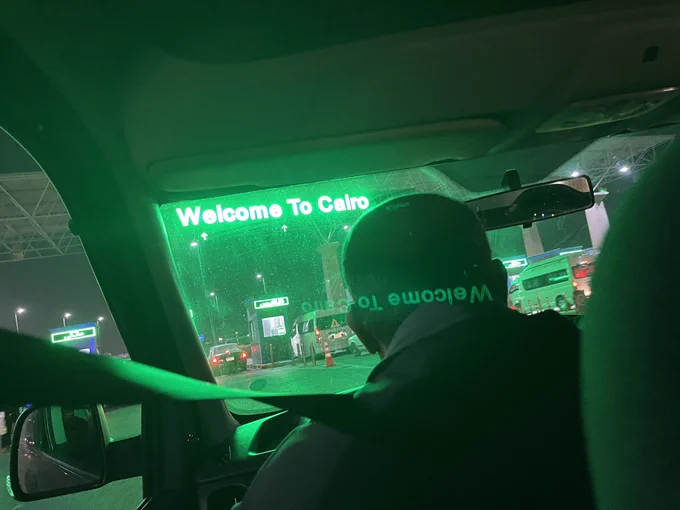 Driving into a checkpoint with a sign saying “welcome to Cairo” in glowing green letters at the top.