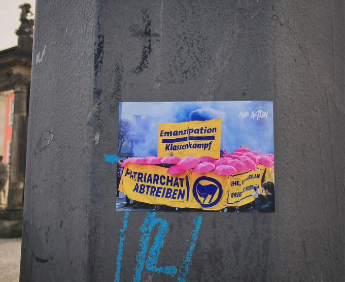 sticker an einem pfeiler einer straßenbahnoberleitung:  es ist ein foto einer demo mit blauem rauch, pinken regenschirmen und gelben bannern mit lila schrift: Emanzipation Klassenkampf PATRIARCHAT ABTREIBEN 

oben rechts ist ein mob action logo