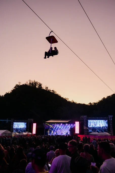 Two people in a chairlift drift through a dusk sky high above a crowd of people watching the Mona Foma stage at the Gorge.