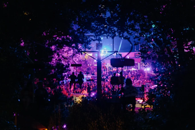 The camera looks through the foliage out from the chairlift entry to the first basin crowd, bathed in coloured lights.