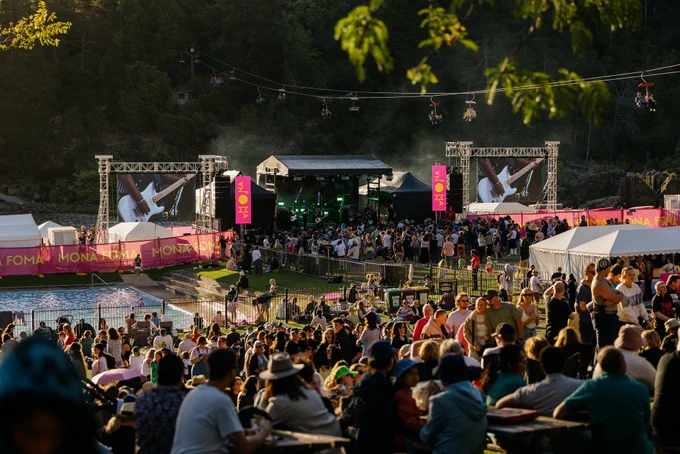 The camera is looking out over lots of people chilling out in the Gorge first basin watching a band on the Mona Foma stage. The Gorge scenic chairlift drifts over the scene with plenty of people enjoying the view from up high.