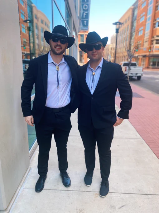 Tyler Halls (left) and Austin Hasen (right), both in black suits and white dress shirts with bolo ties and black cowboy hats, pose on the side walk together with smiles.