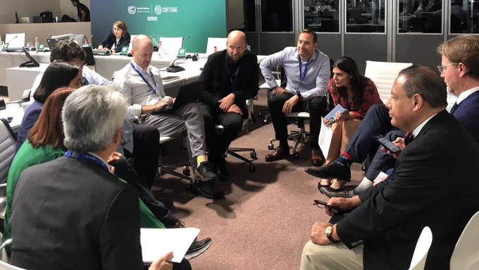 A group of people of various genders and races in suits and other formal office attire in office chairs gathered in a circle, intently discussing a topic