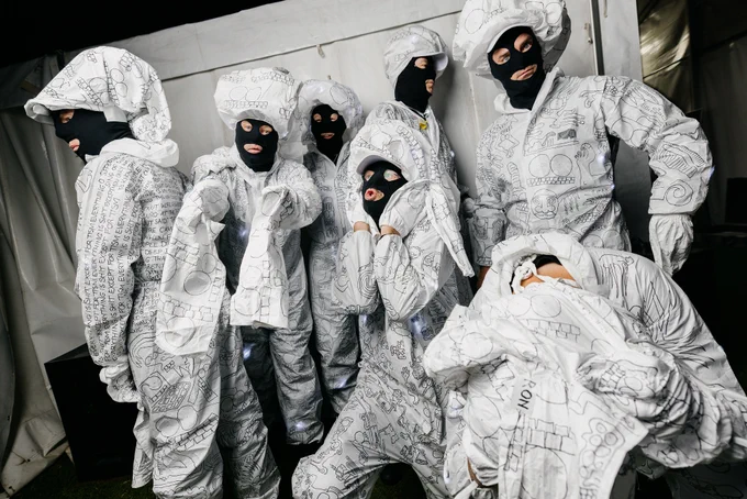 Seven members of the band T.I.S.M posing backstage, dressed in white jumpsuits and black balaclavas. 