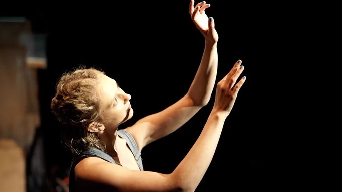 A young woman with blonde curly hair tied in a braid stands on stage. She is wearing blue dungarees and holds her arms up to a light as if it were the sun
