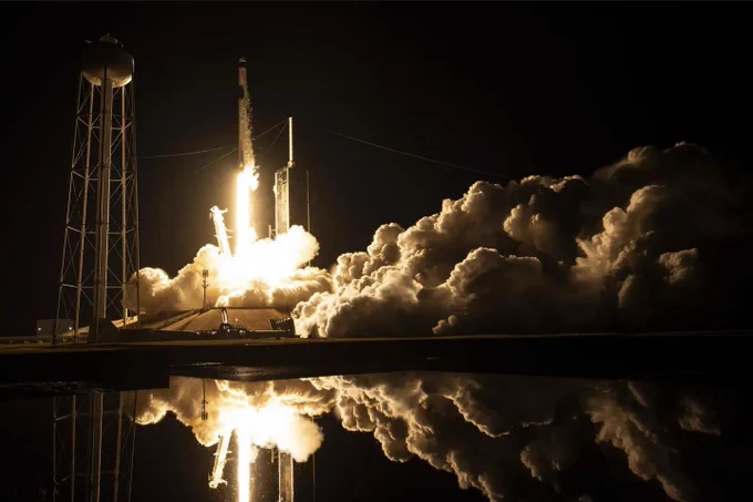 A SpaceX Falcon 9 rocket lifts off into the night sky from NASA's Kennedy Space Center. Flames erupt from beneath the white column of the Falcon, sending plumes across the right half of the image. The launch is partially reflected by a body of water at the bottom of the photo. Credit: SpaceX