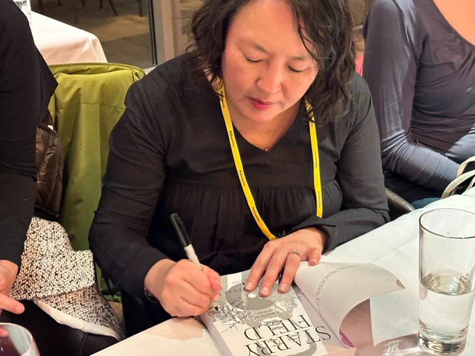 UT alum Margaret Juhae Lee is seen signing her new memoir. She is wearing a black dress with long sleeves and has a yellow lanyard hanging from her neck. The photo is taken from a slightly higher angle from the left side as she holds a sharpie marker to sign the book that is opened, with the pages having “Starry Field” in bold large lettering and “A Memoir of Lost History” in smaller lettering. Image courtesy of Janet Joy Wilson.