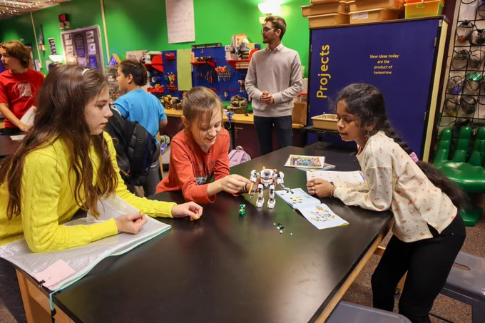 Students engaging in a robotics activity at a classroom table while a teacher supervises in the background.