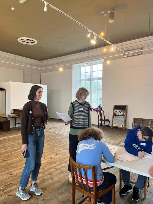 Two workshop leaders stand, smiling and speaking to the participants. The Young People are sat at a table, engrossed in a creative writing task. 