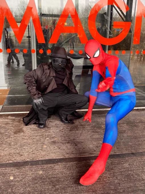 Two figures in costume as Spider-Man noire and classic Spider-Man outside the Magna Centre window - window reads “MAGNA” in big red letters