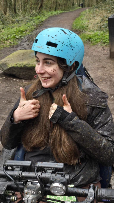 Beth, a woman in her 20s with brown hair, is covered in mud and giving a thumbs up