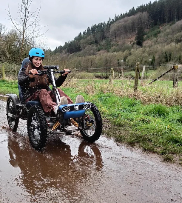 Beth, a young woman with brown hair, is sat in an all terrain wheelchair whizzing along a muddy path 