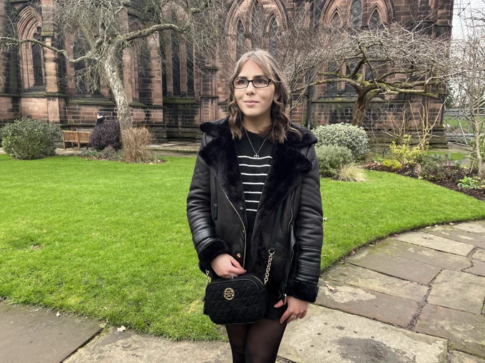 Elin standing on a stone path in front of a large cathedral which has large gothic style windows. The path is bordered by a manicured lawn. She wears a black faux fur lined aviator jacket over a black and white crop jumper and black skort. The outfit is accessorised with a black crossbody bag which has gold hardware, silver jewellery and Elin's trademark black glasses which have diamonds on the side.. Her brown hair is styled into curls and is worn in a half-up, half-down do. She's looking away reflectively to her right.