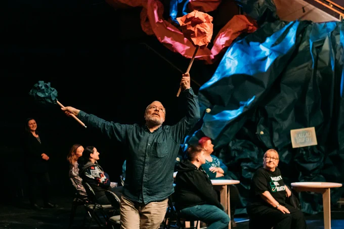 Alex Elliott runs holding two large sticks with blue and orange paper attached to them. A seated audience looks on and there are large paper structures in the background.