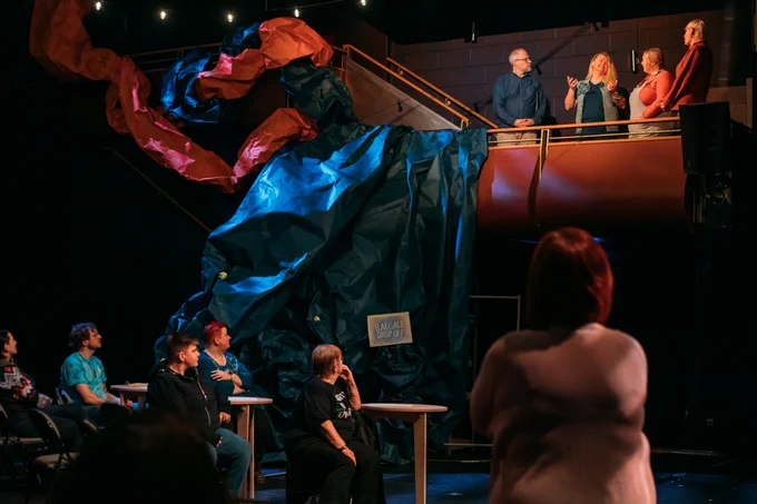 Performers stand on a balcony draped in bright paper, the audience stand and sit on chairs on the ground, looking upwards