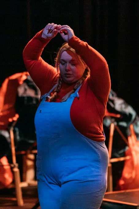 Harrison wearing an orange top and denim dungarees looks down with arms up in the air