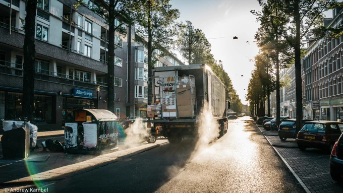 A delivery truck standing on a residential street in dimmed light with delivery items waiting to be unloaded, sorrounded by cars parked mostly on one side of the street and waste containers on the other.