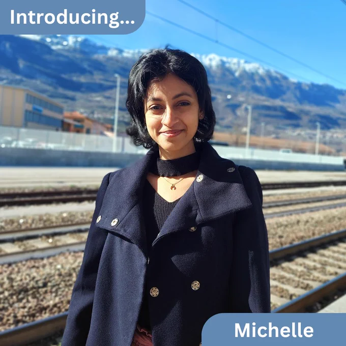 A photograph of Michelle standing outdoors with train tracks and mountains in the background