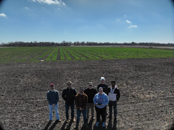 Photo taken from a drone hovering in the air. 7 people pose for a photo in front of a field with plots of wheat.