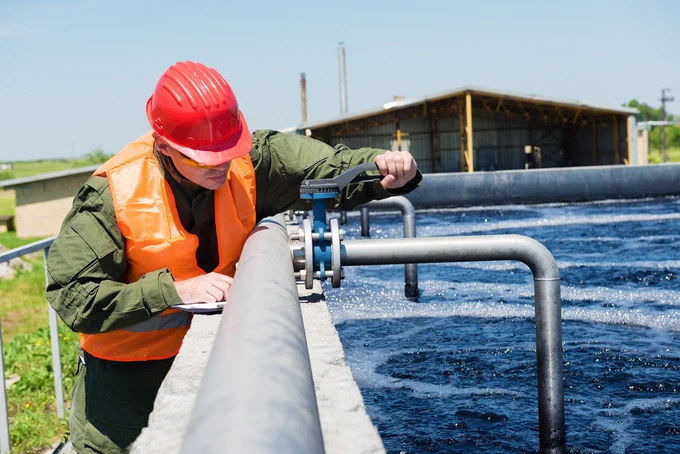 Man in an orange safety vest stands beside an aboveground water storage tank and adjusts a pipe.