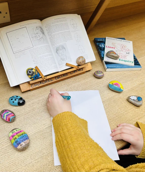Student's arm writing notes from a book in a stand. Colourful rocks are scattered around the desk.