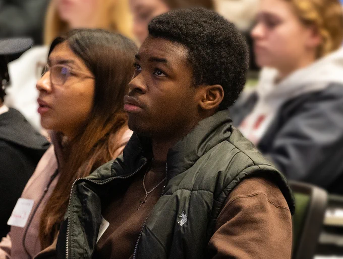 Two students, one with long hair and a sweatshirt and one short, dark, cropped hair look upwards towards the presenters

Two students listen to the opening presentations at the March 2024 College in the Schools event.