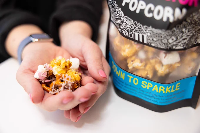 Two hands hold up a handful of Down to Sparkle gourmet popcorn next to a bag of Down to Sparkle.