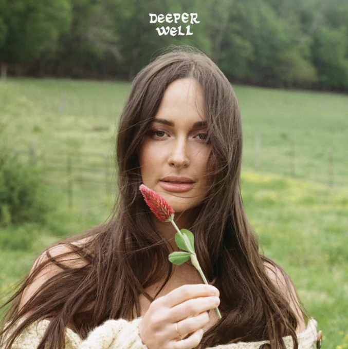 The album cover for Kacey Musgraves’ Deeper Well. Musgraves stands in focus in the center of the frame looking at the camera with a blank look. Behind her is an out of focus grassland with fences along the landscape and trees in the very back. Musgraves is holding a red wildflower with a green stem up to the camera. She is wearing a white off-shoulder sweater and her body is covered by her long brown hair, which is being slightly blown aside by the wind. Above Musgraves is the words “Deeper Well” printed in white in a small playful font. 