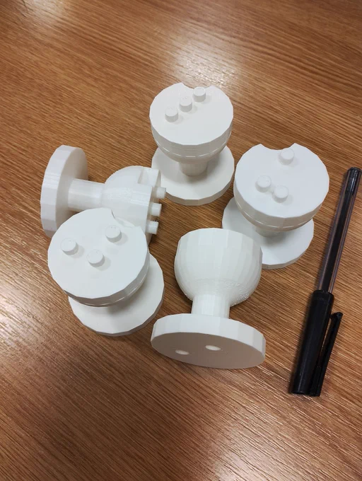5 play dough stampers standing or lying on their side on top of a varnished wooden surface. There is a black biro pen on the right hand side for comparison in size. The base of  each stampers is circular. The holes on the base can be seen. Coming out of the middle of the base is a short stem that widens into a goblet shape. On the top face of the stampers are raised circles depicting the Braille. On the top face is a small semi-circular cut out of the top edge. All the stampers are made of white plastic.
