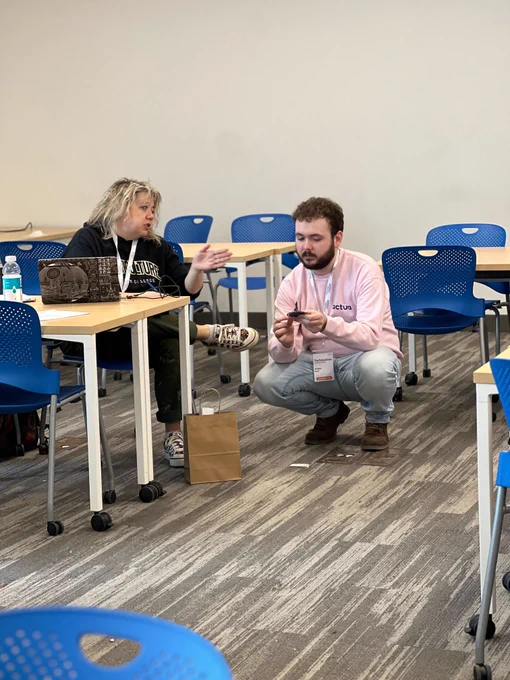 An engaging discussion between two people, one seated at a table with a laptop and the other crouched beside her in a pink "actua" sweater. They appear to be in deep conversation in a classroom with tables arranged for a workshop.
