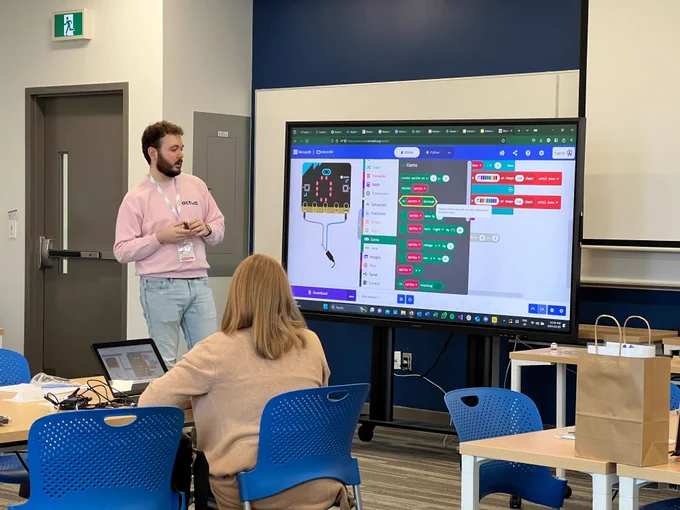 A presenter in a pink sweater labeled "actua" is standing in front of a large display screen showing a colorful coding interface. The audience, seen from behind, is attentively watching the presentation in a classroom setup with laptops open on the tables.