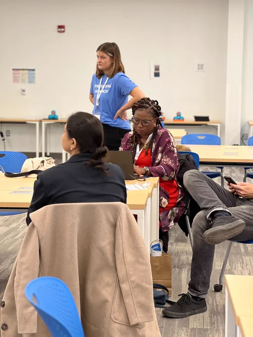 A workshop facilitator in a blue "Engineering Outreach" t-shirt is standing, observing a participant who is focused on a laptop screen. The setting is a classroom environment with blue chairs and tables, where some educational interaction is taking place.