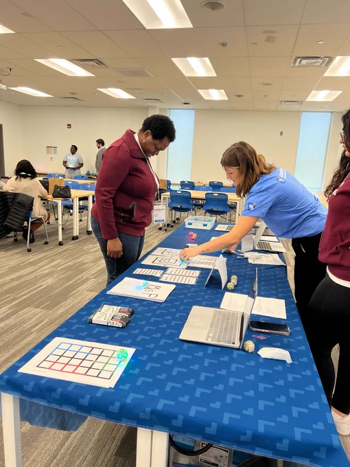 Two people are interacting at a workshop with a blue tablecloth. One is intently observing a robotics activity, while the other, in a blue t-shirt, is assisting and pointing towards a robotic device next to a laptop, indicating a hands-on learning environment.