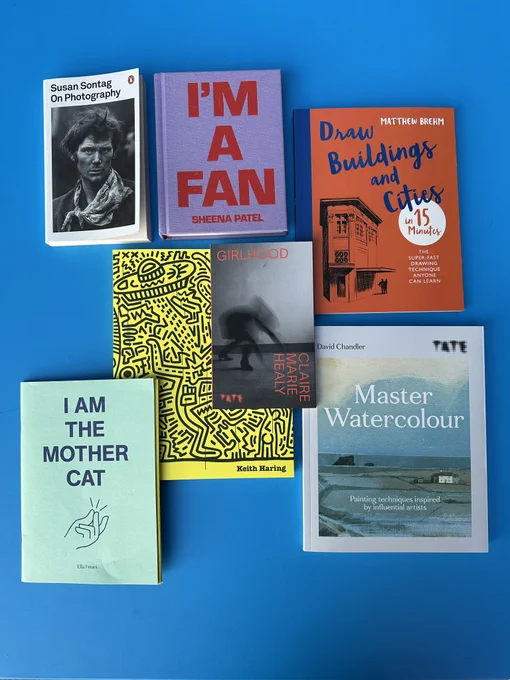 A photograph of 7 books displayed on a blue background. The books are: On Photography by Susan Sontag, I'm a Fan by Sheena Patel, Drawing Buildings and Cities in 15 minutes by Matthew Brehm, Master Watercolour by David Chandler, Girlhood by Claire Marie Healy, Keith Haring by Darren Pih and I am the Mother Cat by Ella Frears.