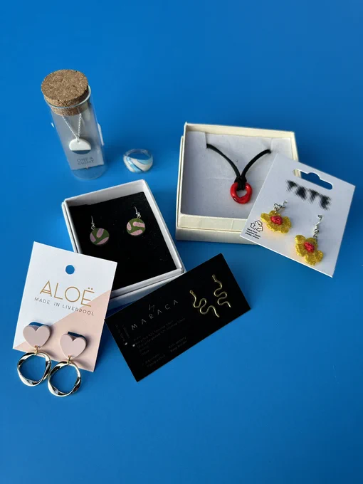 A photograph of a selection of jewellery displayed on a blue background. It includes earrings by One & Eight, ring and necklace by Roslyn, earrings by Aloë, earrings by Tate and earrings by Jemma Marston.