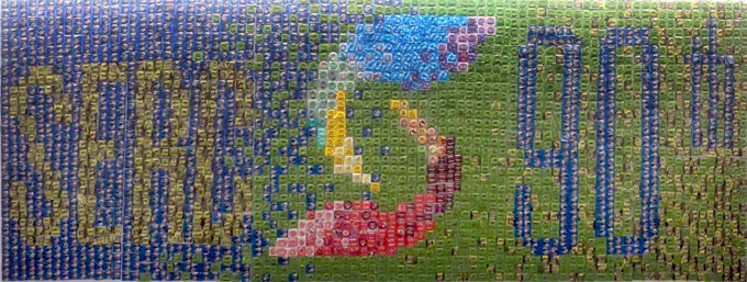 A long narrow photo of a wall mural made of condoms. On the left side of the mural it says "SERC" in green condoms, on the right side of the mural it says "90th" in blue condoms. In the middle there is the rainbow "S" from SERC's logo. The background is a gradient of blue to green, also made of condoms. 