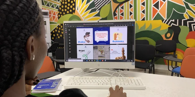 A woman at a computer is looking at images of educational language content with a colorful mural in the background.