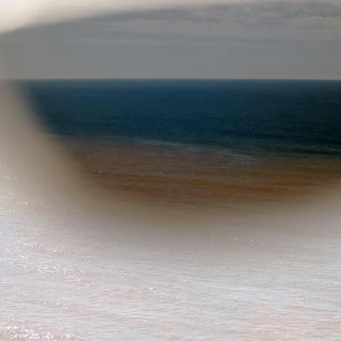 A slightly blurry scene of deep blue water turning brown as it meets the shore, with a flat clear horizon behind.