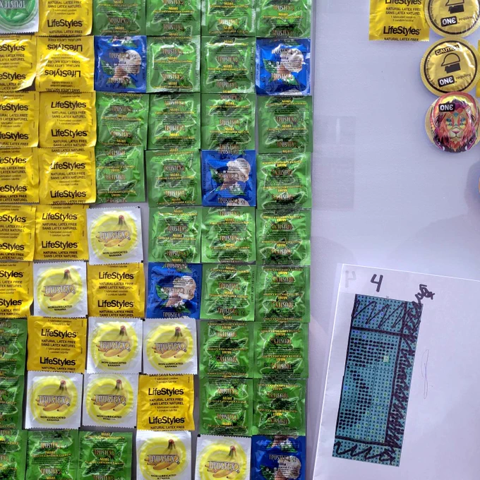 A close up picture of condoms taped to a white wall. There is a picture taped to the wall next to the condoms, that shows a pattern for the design. 