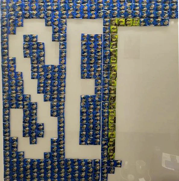 A picture of the condom mural in progress. Blue condoms are taped to the wall, and blank spaces reveal the white wall and spell "S" and "E".