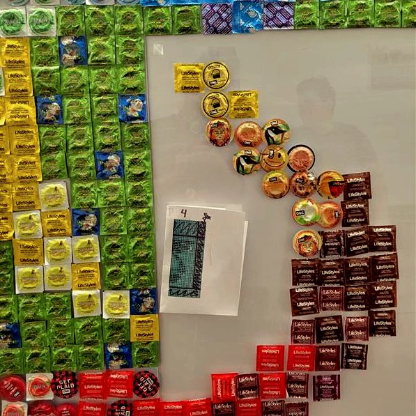 A close up picture of condoms taped to a white wall. There is a picture taped to the wall next to the condoms, that shows a pattern for the design. 