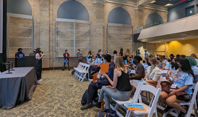 A photograph of a group of people, seated and taking up the right-hand side of the photo, listening attentively to the speaker. The speaker holds a microphone and stands on the left-hand side of the photo. Upcoming speakers are lined up against the far wall of the large conference room. 